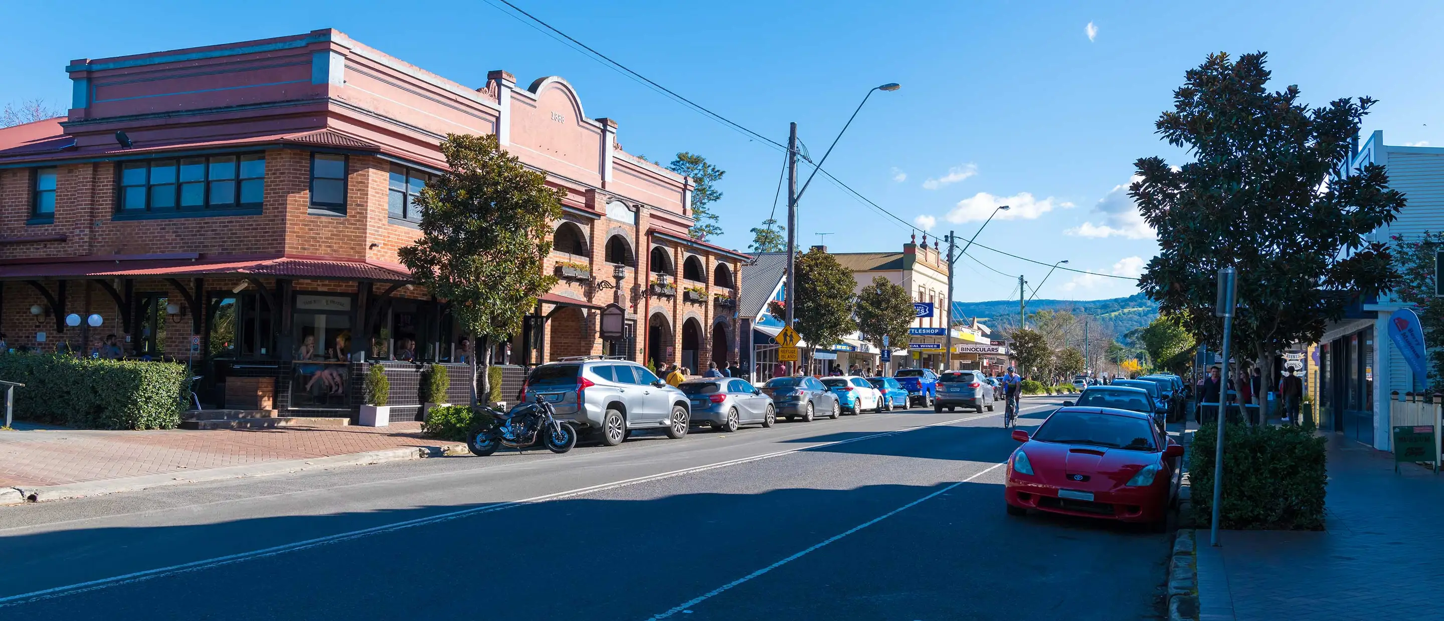 Sunny main street lined with heritage shopfronts, a brick hotel with arched verandahs on the corner and green hills behind